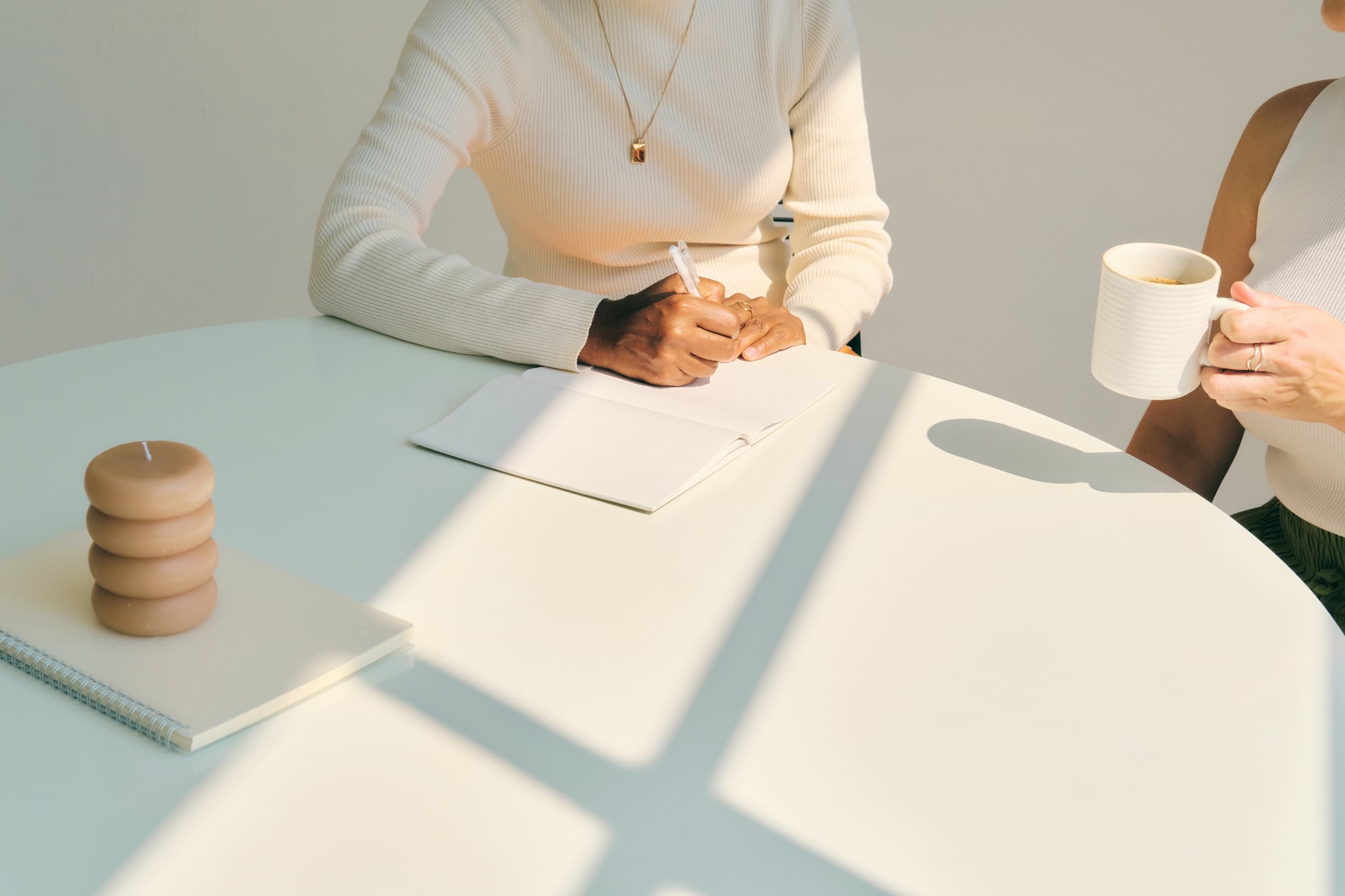 Two people in conversation at a sunlit table, one writing in a notebook while the other holds a cup